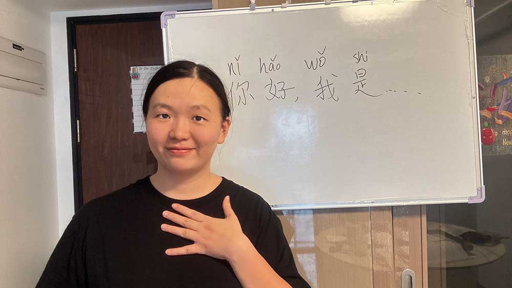 Candice, a Mandarin tutor, smiling in front of a whiteboard with a Chinese greeting and pinyin.